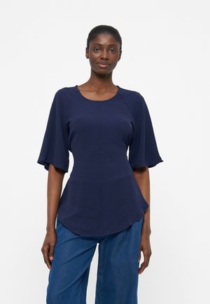 Kobieta stojąca, ubrana w granatową bluzkę z szerokimi rękawami do łokcia oraz niebieskie, szerokie spodnie z wysokim stanem na jednolitym tle.