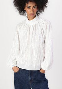 Hvit bluse med teksturerte bladlignende mønstre, høy hals og rynkede mansjetter. Kombinert med mørkeblue jeans for kontrast.