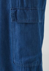 Blå denimstoff med en jevn tekstur og lett glans, med en lappedeksel og kontrasterende sømmer langs sømmene.