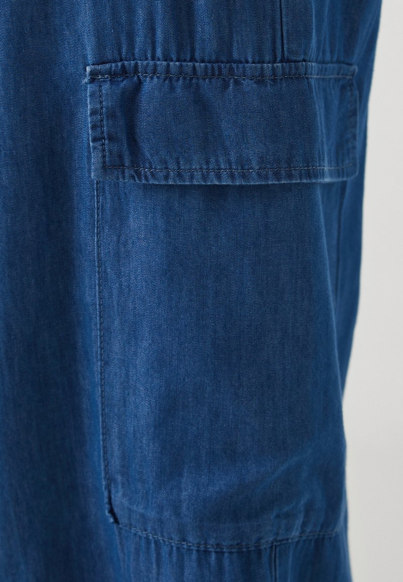 Tissu en denim bleu avec une texture lisse et un léger éclat, comportant une poche plaquée et des coutures contrastées le long des seams.