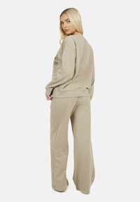 Vrouw met lang blond haar, gekleed in een beige oversized sweatshirt en wijde broek, kijkt over haar schouder tegen een effen witte achtergrond.