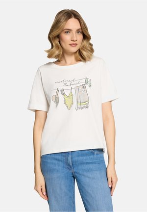 Frau trägt ein weißes T-Shirt mit einer Illustration von Strandmode und dem Text "Meeting am Strand", kombiniert mit blauen Jeans, steht vor weißem Hintergrund.