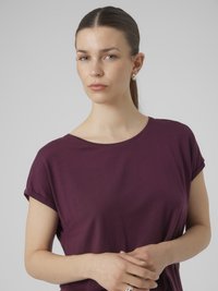 Kvinne med rett brunt hår satt opp, iført sølv hoop øreringer og en kortermet lilla skjorte, med hendene løst sammenfoldet.
