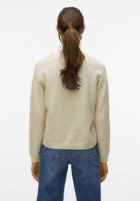 Maglione beige chiaro con una trama morbida, caratterizzato da un collo rotondo e polsini a coste, abbinato a jeans in denim blu con tasche posteriori.