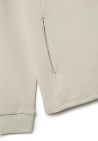 Tejido de sudadera color beis claro con un bolsillo lateral con cremallera, puños acanalados y una textura suave con detalles de costura limpios.