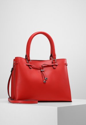 Červená kožená kabelka s dvojitými držadly, dekorativním uvázaným páskem a odnímatelným ramenním popruhem na bílém podstavci proti světle šedému pozadí.