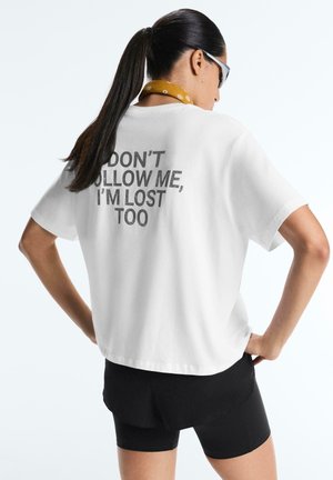 Žena s copom, ktorá nosí biele tričko s nápisom "NEDEPTAJ MA, AJ JA SOM STRATENÁ," čierne šortky, slnečné okuliare a žltý šál okolo krku.