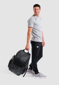 Czarna sportowa plecak z siateczkowymi akcentami, z wieloma przegrodami; mężczyzna w szarym T-shircie i czarnych spodniach trzyma go w neutralnej pozie.