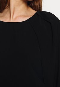 Vêtement noir avec un col rond et des épaules froncées. Le tissu a une texture douce et il y a des plis subtils le long des coutures.