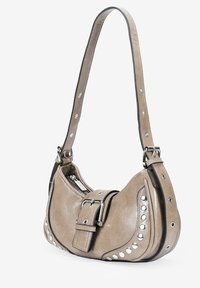Bolso de cuero marrón claro con forma curvada, tachuelas plateadas a lo largo del costado y un detalle de hebilla prominente. Incluye una correa de hombro desmontable.