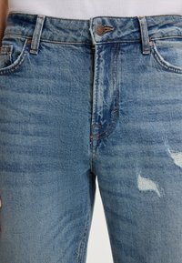 Niebieskie dżinsy z denimu z przednim guzikiem, szlufkami na pasek, widocznym szwem i przetarciami na prawej udzie, noszone z białym topem.