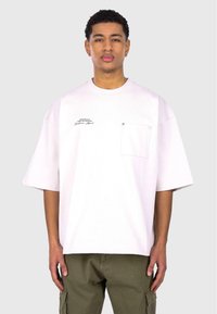 White oversized t-shirt with short sleeves, a front pocket, and printed text on the left side. Worn with olive green cargo pants.