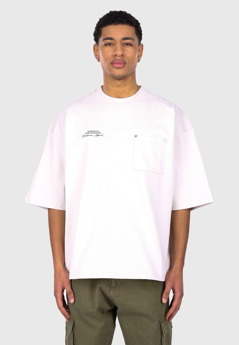 White oversized t-shirt with short sleeves, a front pocket, and printed text on the left side. Worn with olive green cargo pants.
