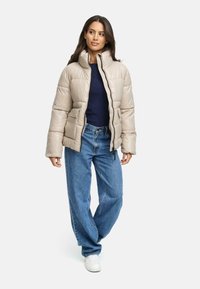 Beige pufferjack met een hoge kraag en rits aan de voorkant, met een doorgestikt ontwerp en zijzakken, gecombineerd met blauwe, loszittende jeans en witte sneakers.