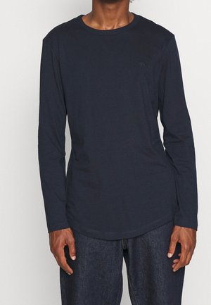 Personne portant un t-shirt uni à manches longues bleu marine foncé avec un col rond et un jean bleu foncé, debout devant un fond clair.