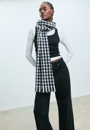 Schwarz-weißer karierter Schal mit fransigen Kanten, getragen über einem schwarzen Tanktop und einem weißen Langarmshirt. Dunkle weite Hosen.