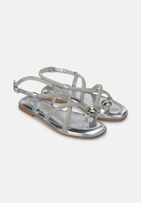 Zilveren sandalen met kruislings straps, met een metallic glans, textuur op de straps en een gespsluiting. De voetbed is plat met een lichte zool.