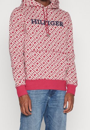 Osoba nosząca czerwoną i białą wzorzystą bluzę z kapturem z napisem "HILFIGER" i niebieskie dżinsy, pokazana od ramion do połowy uda na jednolitym tle.