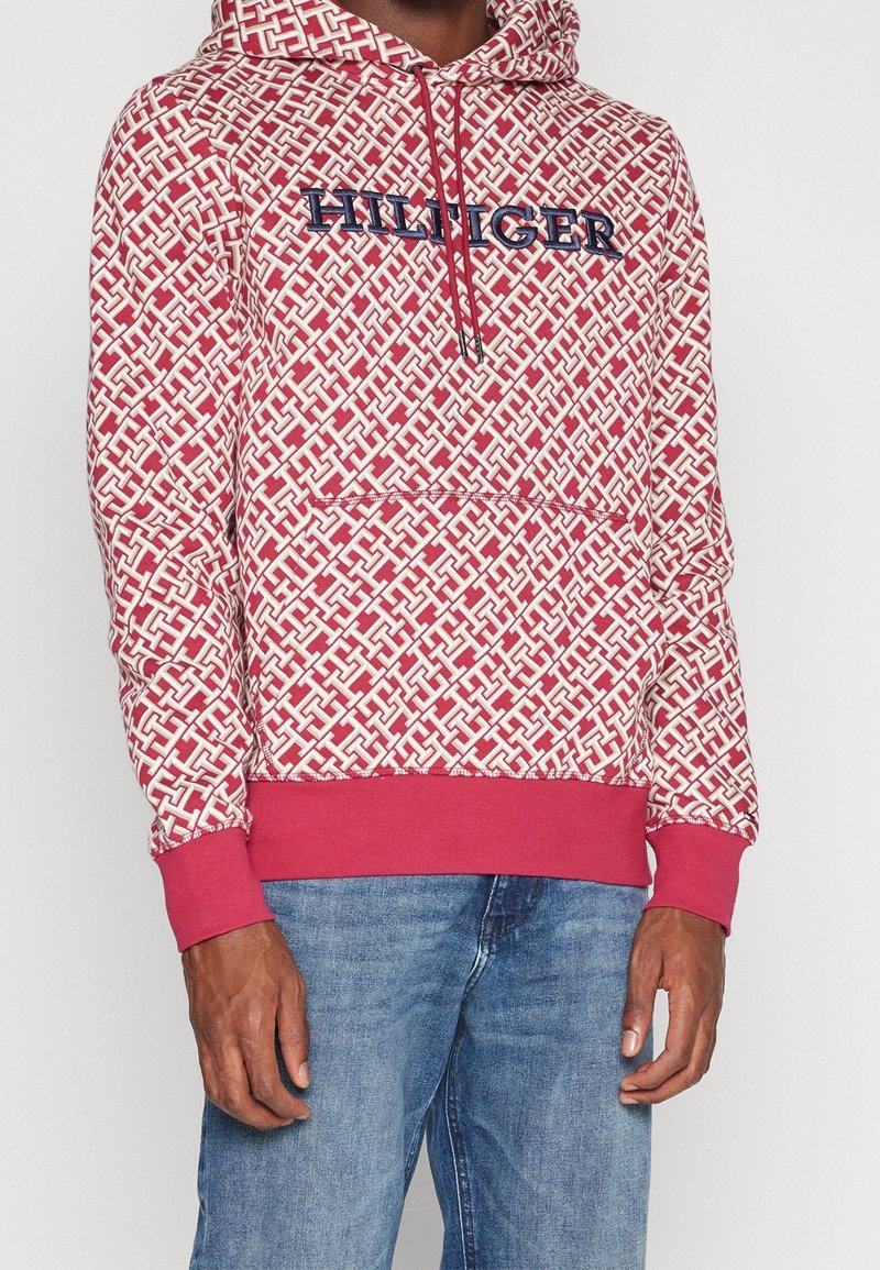Personne portant un sweat à capuche rouge et blanc à motifs avec le texte "HILFIGER" et un jean bleu, visible des épaules jusqu’au milieu des cuisses sur un fond uni.
