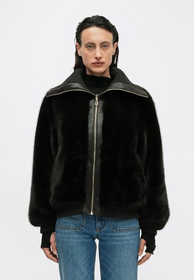 Giacca in pelliccia sintetica nera con zip frontale, collo strutturato e polsini elasticizzati; abbinata a jeans blu e a un dolcevita nero sotto.