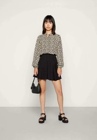 Blusa preta com padrão e mangas bufantes, combinada com uma saia preta de corte evasê. A modelo segura uma pequena mala preta e usa sandálias de plataforma pretas.