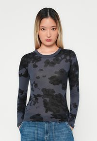 Camisa de manga larga de color gris oscuro con un patrón de camuflaje negro, cuello redondo, diseño ajustado y textura suave. Combinada con jeans de mezclilla azul.