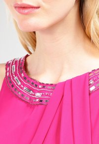 Närbild av en person som bär en ljusrosa klänning med en pärlbesatt och spegelbelagd halsringning, som visar läppar, haka och hals.