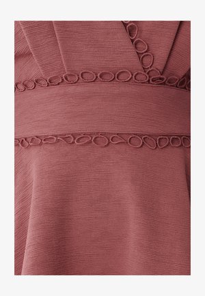 Frambozenkleurige stof met een gestructureerd oppervlak. Heeft gestikte ronde accentueringen langs de taille, wat een subtiele designelement creëert.