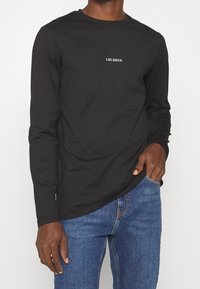 Langärmliges schwarzes T-Shirt aus Baumwolle mit Rundhalsausschnitt und weißem "LES DEUX"-Logo auf der Brust. Glatte Textur mit taillierter Passform.