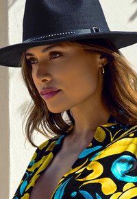Black felt hat features a decorative band with studs. The garment has a vibrant floral pattern in yellow and blue against a black background.