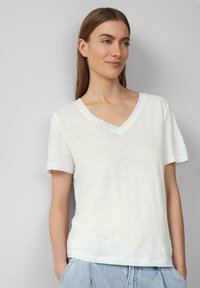 Biała koszulka T-shirt z dekoltem V i krótkimi rękawami, wykonana z teksturowanej tkaniny, o luźnym kroju. Noszona z jasnoniebieskimi szortami dżinsowymi.