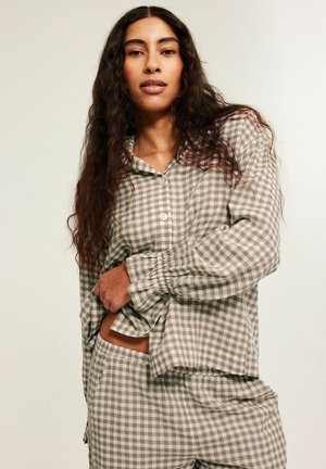 Vrouw met lang krullend haar, kleed zich in een beige-wit geruit overhemd met lange mouwen en bijpassende broek, tegen een effen achtergrond.