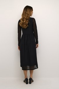 Robe maxi noire avec un corsage ajusté et une jupe évasée, dotée de manches longues transparentes et d'un détail de taille froncé. Des chaussures à talons noires complètent le look.