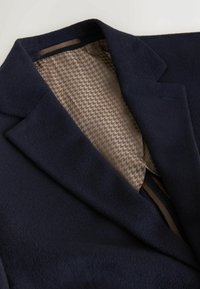 Mörkblå ullblazer med klyvna krage och foder i hounds-tooth mönster i beige. Har rena linjer och mjuk textur.