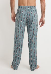 Heren pyjamabroeken van een teal stof, met een patroon van nutcracker figuren. Elastische tailleband, rechte snit, lichtgewicht materiaal.