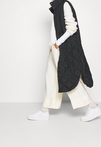 Person som går i vita vida byxor, matchande tröja, lång svart vadderad väst och vita sneakers mot en enfärgad bakgrund.