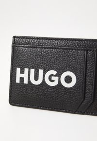 Portafoglio in pelle nera con texture, caratterizzato da più scomparti e audace logo bianco "HUGO" sulla parte anteriore. Design minimalista, forma compatta.
