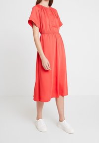 Robe midi corail avec manches courtes, taille élastique et détails de boutons. Tissu lisse avec une silhouette fluide, associée à des baskets blanches.