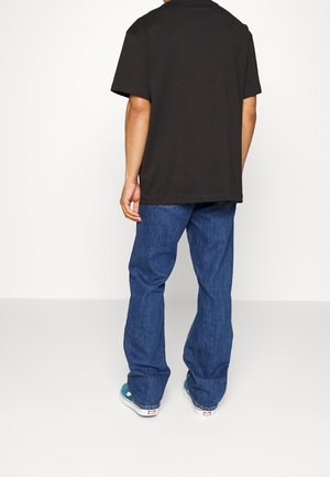 Person trägt ein schwarzes Kurzarmshirt, blaue Jeans und türkisfarbene Turnschuhe und steht mit dem Rücken zu uns vor einem schlichten weißen Hintergrund.