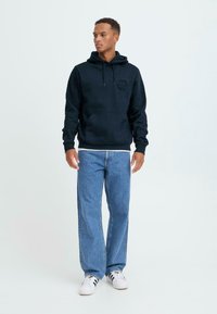 Svart hettegensere med en frontlomme og teksturert logo, sammen med lyseblå rettbukser og hvite sneakers med svarte striper.