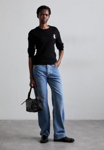 Maglione nero a coste con una patch di un orso, jeans a gamba larga blu chiaro, tiene una borsa nera e indossa scarpe nere. Sfondo minimalista.