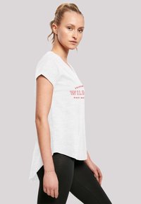 Balta trumpa rankovė t-shirt su apvaliu apvadu, su raudonu teksto spaudiniu. Marškiniai vilkimi virš juodų leginsų, demonstruojantys kasdienį stilių.