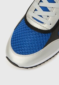 Kék hálós sportcipő ezüst szintetikus ráhímzésekkel, fekete díszítésekkel és fehér fűzőkkel. Texturált felülettel és lekerekített orr-résszel rendelkezik.