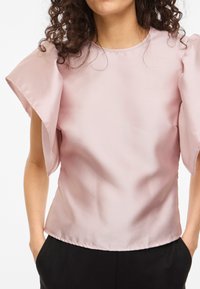 Naine kannab heleroosat satiinist pluusi suurte lehvidega varrukatega ja musti pükse, käed taskutes, nähtavad lokkis tumedad juuksed.