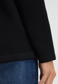 Czarny sweter z textured o splocie w kratkę, z prążkowanym dołem i mankietami, zestawiony z dżinsami. Zbliżenie na rękaw i materiał.