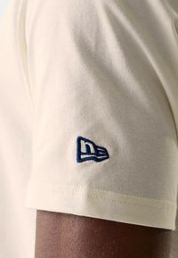 Camiseta de algodón color crema con un logo bordado azul en la manga izquierda, de manga corta y con una textura suave y lisa.