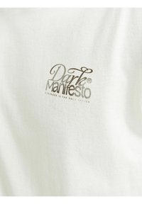 Bijela pamučna majica sa printed logom "Dark Manifesto" u srebrnoj boji i tagline-om "Stronger Is The Only Option" ispod.