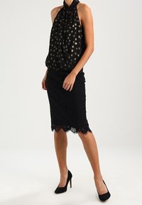 Femme portant un haut noir sans manches à col licou avec des pois dorés et une jupe noire en dentelle jusqu'aux genoux, accompagnée de talons hauts.