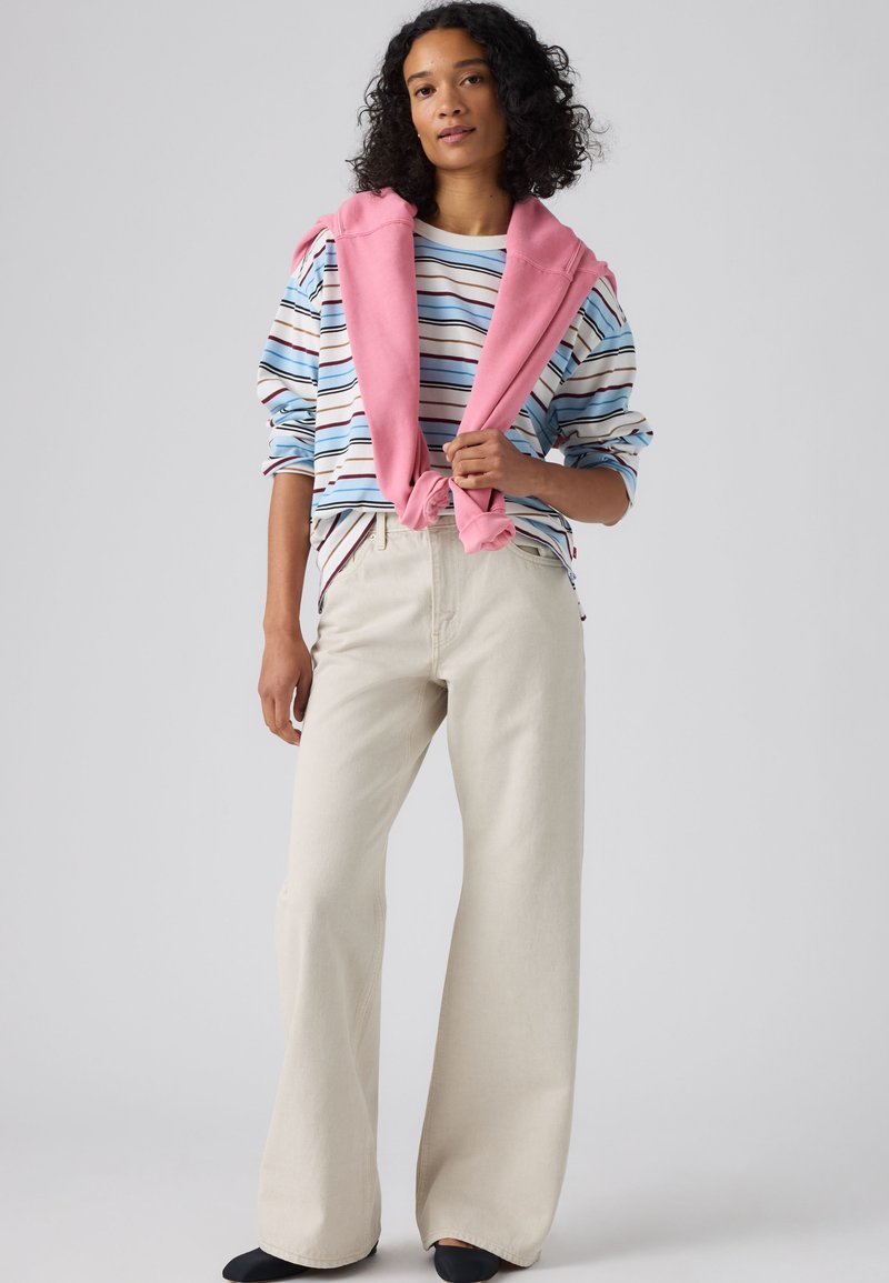 Femme aux cheveux bouclés portant une chemise rayée et un pantalon ample beige, tenant un sweat-shirt rose drapé sur ses épaules.