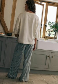 Světlý, volný top s širokým výstřihem, v kombinaci s modrými pruhovanými pyžamovými kalhotami, které mají malé, hravé ilustrace.
